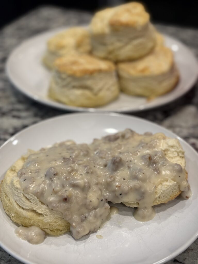 Golden brown buttermilk biscuits served with creamy homemade sausage gravy in a skillet and ladled over warm biscuits on a plate—classic Southern-style breakfast comfort food