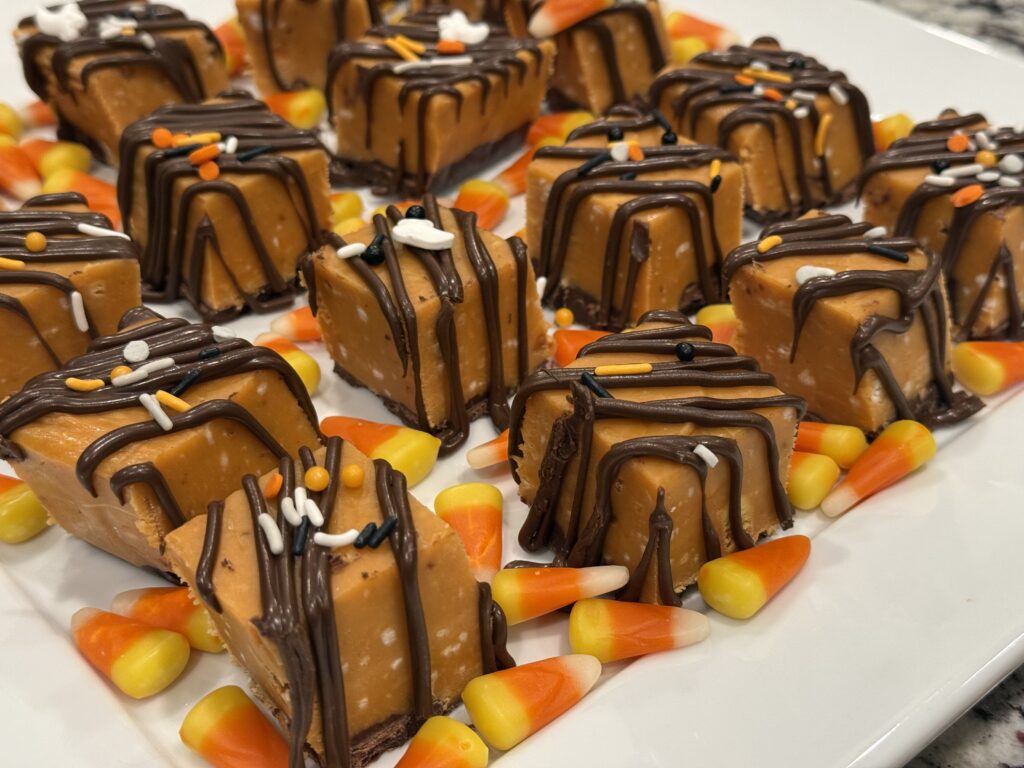 Close-up of square pieces of orange peanut butter fudge drizzled with melted chocolate and Halloween sprinkles, arranged on a platter with candy corn.