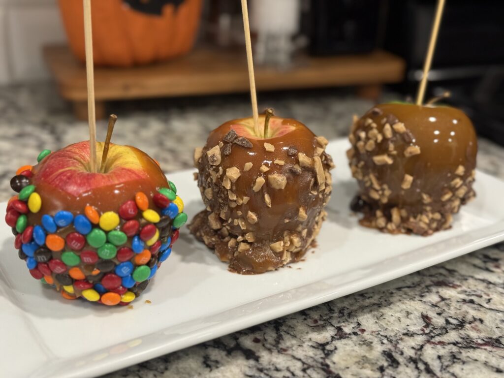 Three homemade caramel apples on a white tray, coated with glossy caramel and rolled in M&Ms and toffee bits, with a smiling jack-o’-lantern decoration in the background.