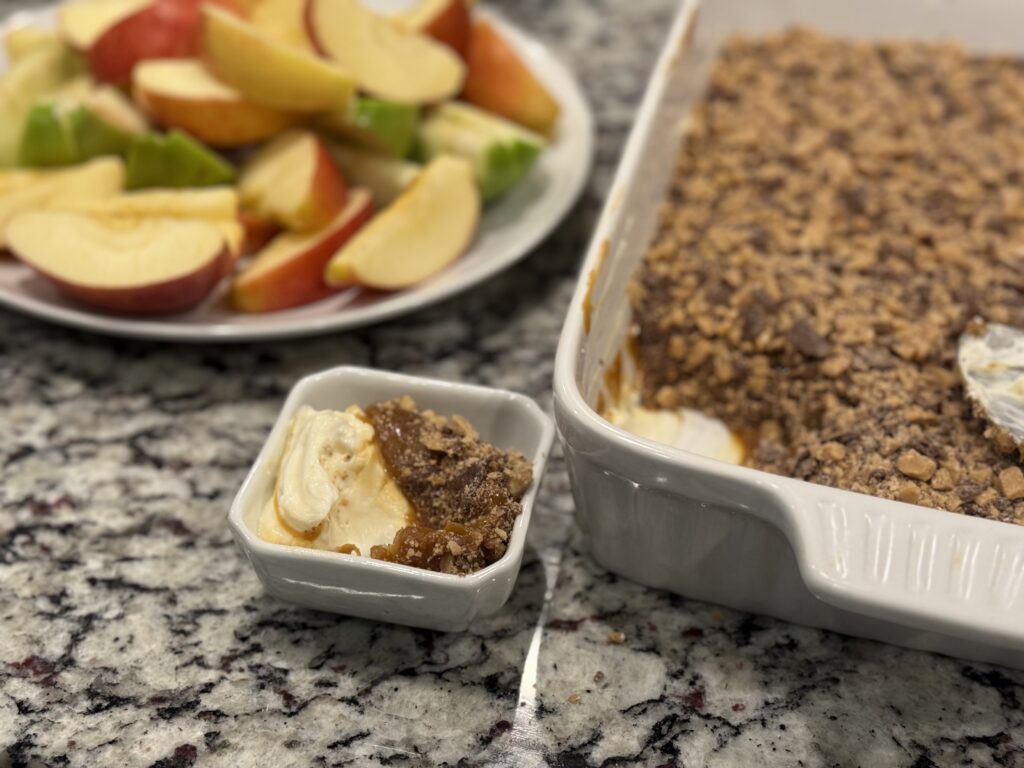 Caramel apple dip in a 9x13 dish with layers of whipped cream cheese, thick caramel, and toffee bits, surrounded by sliced apples for dipping.