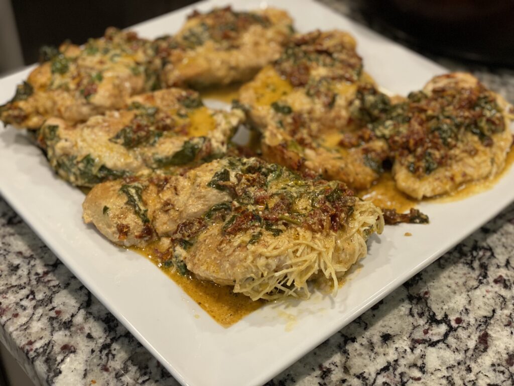 Close-up of creamy Tuscan chicken on a white platter, topped with sun-dried tomatoes, spinach, and melted Parmesan in a rich golden sauce.