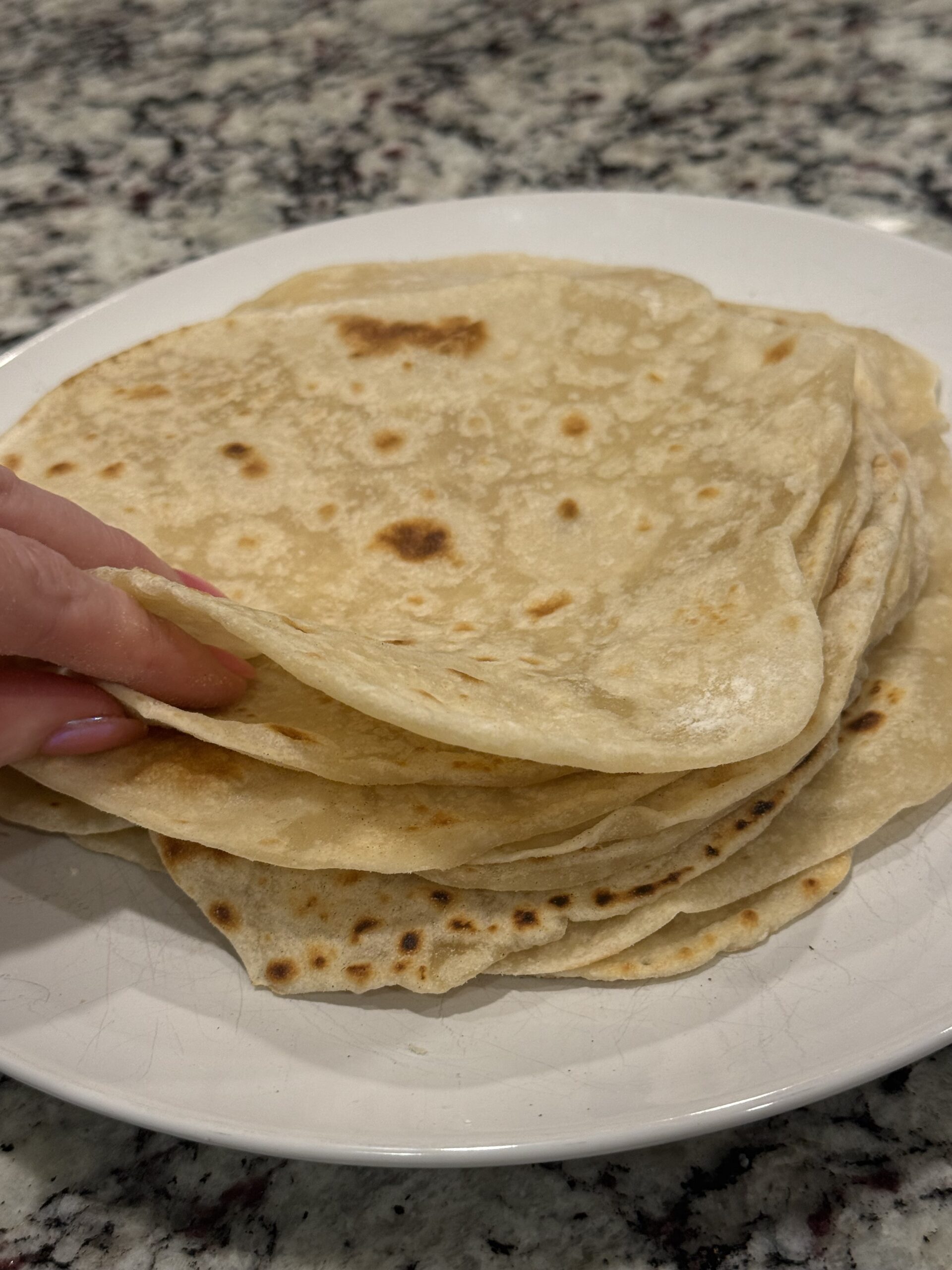 A stack of homemade flour tortillas piled on a white plate. The tortillas are soft and thin with golden brown spots from cooking on a skillet. The edges are slightly uneven, highlighting their homemade appearance. The tortillas are stacked high to showcase their soft texture and flexibility, making them perfect for tacos, burritos, quesadillas, or wraps.