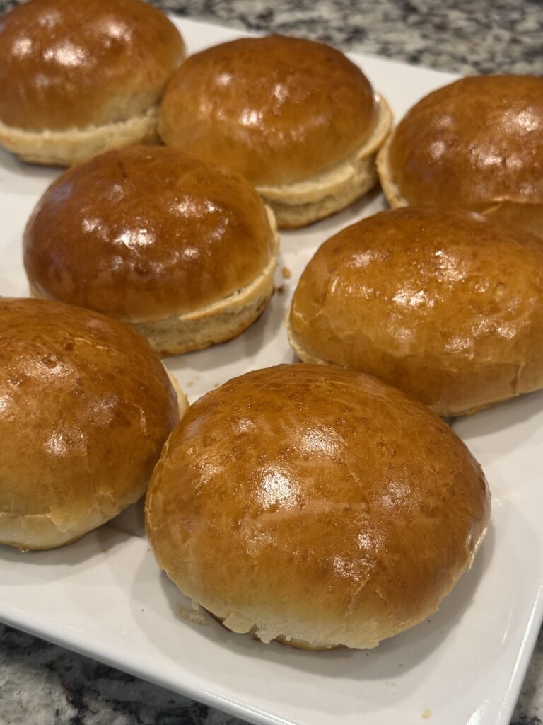 Freshly baked homemade burger buns with shiny golden brown tops arranged on a white serving platter.