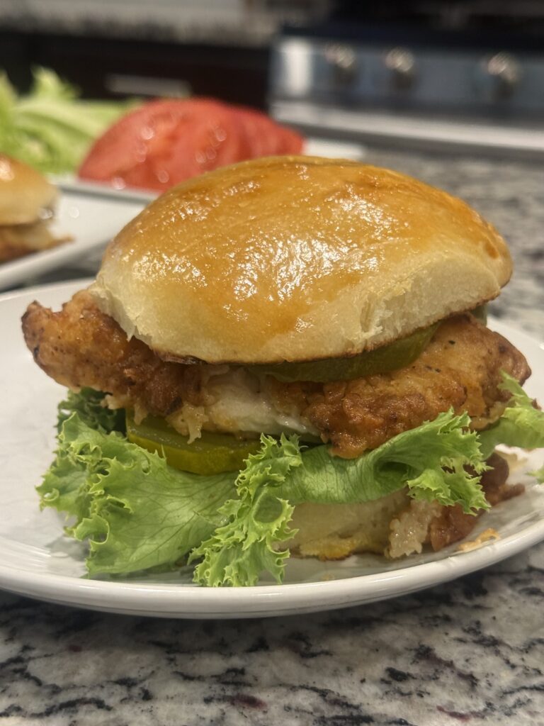 Golden crispy fried chicken sandwich on a homemade burger bun with lettuce, tomato, pickles, and mayonnaise served on a white plate.